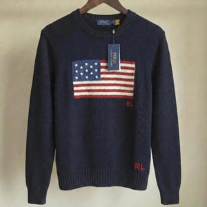 May include: A navy blue long-sleeve crewneck sweater featuring a knitted American flag design on the chest. The sweater includes red embroidered RL initials below the flag and a Polo Ralph Lauren brand tag attached to the collar with a plastic fastener.