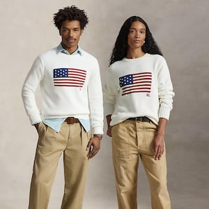 May include: Two white long-sleeved knit sweaters featuring a prominent American flag graphic on the chest with the letters RL embroidered in red. The sweaters are paired with light blue collared shirts and khaki trousers with brown belts.