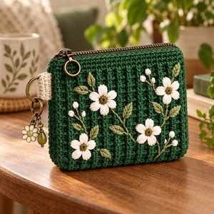 Può includere: Una pochette all'uncinetto verde scuro con cerniera, decorata con fiori bianchi ricamati e foglie verdi. La pochette presenta una cerniera in metallo color bronzo e un pendente laterale con una margherita e una perlina verde.