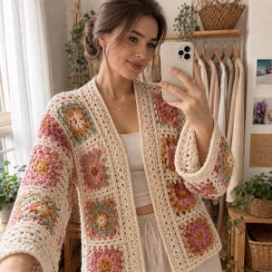 May include: A long-sleeved cardigan made of cream-colored yarn featuring a patchwork of square crochet motifs in shades of pink, yellow, and sage green. The garment has an open front design with a wide border and a relaxed fit throughout the body.