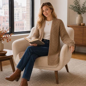 May include: A long-sleeved, open-front cardigan in a textured tan knit fabric, worn over a plain white t-shirt and dark blue denim jeans. The cardigan features a ribbed hem and cuffs, providing a layered look with a simple gold pendant necklace.