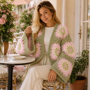 May include: A crochet cardigan featuring a sage green base with large, square floral motifs in shades of light pink, sage green, and cream. The garment has wide sleeves, an open front with thin tie closures, and a textured, fuzzy yarn finish.