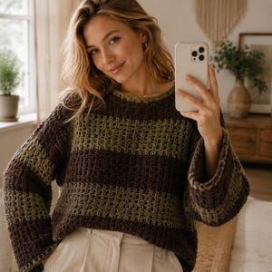 May include: A long-sleeved crochet jumper featuring horizontal stripes in alternating shades of dark brown and olive green. The garment has a round neckline and a loose, textured stitch pattern throughout, paired with light-coloured trousers.