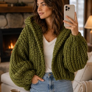 May include: A chunky knit olive green cardigan with a hood and long sleeves, worn over a plain white v-neck t-shirt and light blue denim jeans. The garment features a thick, textured yarn construction with a relaxed fit and open front design.