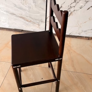 May include: A dark brown wooden bar stool featuring a square seat and a backrest with two scalloped horizontal slats. The stool has four cylindrical legs connected by a footrest, with visible light-colored joinery details at the connection points.