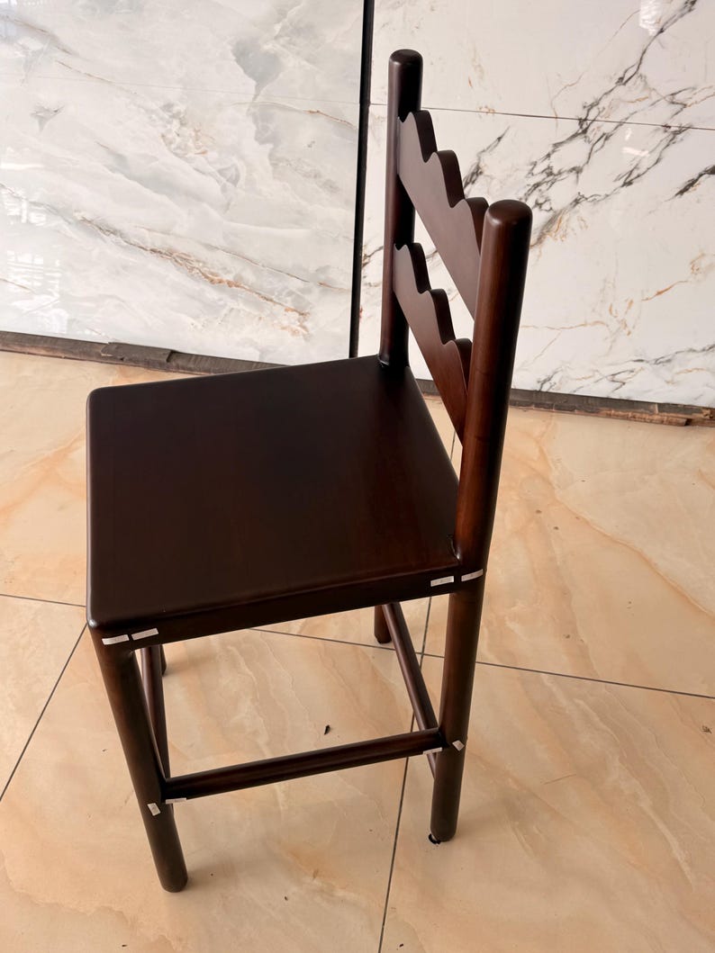 May include: A dark brown wooden bar stool featuring a square seat and a backrest with two scalloped horizontal slats. The stool has four cylindrical legs connected by a footrest, with visible light-colored joinery details at the connection points.