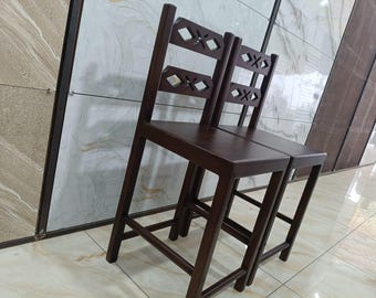 May include: Two dark brown wooden bar stools with square seats and straight legs. The backrests feature a horizontal slat design with two diamond-shaped cutouts in each. The stools have a smooth, dark finish and a simple, functional construction.