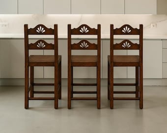 May include: Three dark brown wooden bar stools with square seats and four legs connected by horizontal stretchers. Each stool features a high backrest with two horizontal slats, each containing a decorative cutout shaped like a fan.