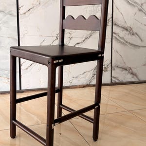 May include: A dark brown wooden bar stool featuring a square seat and a backrest composed of two horizontal slats with a scalloped, wavy edge design. The stool has four straight legs connected by horizontal support bars on all four sides.