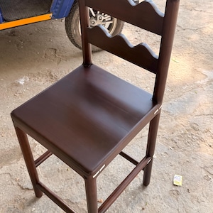 May include: A dark brown wooden bar stool featuring a square seat and a backrest with two horizontal slats, each designed with a scalloped, wavy top edge. The stool is supported by four straight legs connected by horizontal rungs for stability.