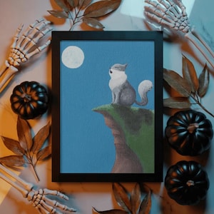 May include: A framed rectangular print featuring a painting of a grey and white cat sitting on a green cliff edge under a full moon against a blue sky. The artwork is displayed in a simple black frame, capturing a nocturnal scene with a feline.
