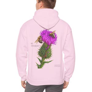 Könnte beinhalten: Ein hellrosa Kapuzenpullover mit einem Aufdruck auf der Rückseite, der eine violette Distelblüte mit zwei Bienen zeigt. Der Text "BEE DIFFERENT" steht links neben der Blume, und "LAURASPRICK FOTOGRAFIE" ist in kleinerer Schrift darunter gedruckt.