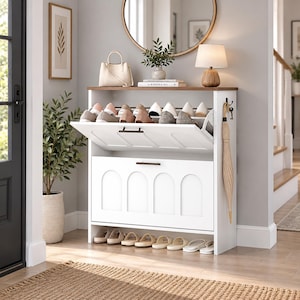 May include: A white shoe storage cabinet with a wood-tone top surface, featuring two tilt-out compartments with arched panel detailing and a bottom open shelf. The unit includes a side hook holding a beige umbrella and dark metal hardware handles.