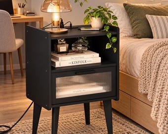 May include: A black bedside table with four tapered legs, an open storage shelf, and a single drawer featuring a ribbed glass front. The top holds a gold-toned mushroom lamp, a small potted plant, and two books titled The Art of Home and The KINFOLK TRAVEL.