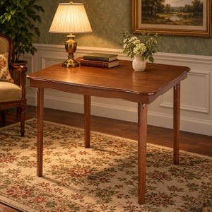 May include: A square wooden table with a warm brown finish and slightly curved edges. The table features four straight legs and a smooth surface. It is positioned in a room with a lamp, two stacked books, and a small vase of white flowers on top.