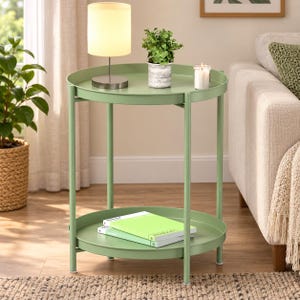 May include: A sage green metal side table with two circular tiers and a raised edge on each tray. The table features four thin vertical legs and a smooth finish. Two stacked books with light green covers rest on the bottom shelf of the table.