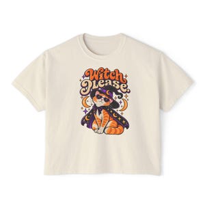 May include: A cream-colored short-sleeve t-shirt featuring a graphic of an orange tabby cat wearing a black witch hat, a purple cape, and sunglasses. Above the cat, the text reads "Witch Please." in orange and purple stylized lettering.