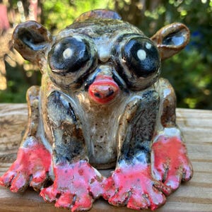 May include: A ceramic figurine of a tarsier with large, dark, glossy eyes, a small red nose, and prominent ears. The body is glazed in mottled shades of brown and grey, while the hands and feet are painted in a vibrant, glossy red finish.