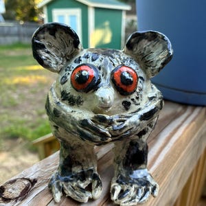 May include: A hand-sculpted ceramic figurine of a creature with large, round, bright orange eyes and a glossy, off-white body covered in irregular black splotches. The figure has prominent ears, short arms held across its chest, and clawed feet.
