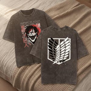 Puede incluir: Una camiseta gris carbón con efecto lavado al ácido que presenta un gráfico de un personaje con cabello oscuro y máscara en el frente, junto con texto japonés y el texto SHINGEKI NO KYOJIN. La espalda muestra un gran emblema de alas blanco y negro.
