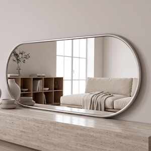 May include: A large, horizontal, oval-shaped wall mirror with a thin, smooth, silver-toned metal frame. The reflective surface shows a room interior with a beige sofa, a wooden shelving unit, and decorative ceramic vases.