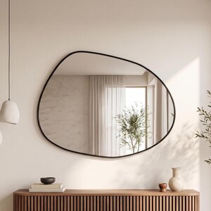 May include: A wall-mounted mirror with an irregular, organic, pebble-like shape and a thin black frame. The reflective surface shows a room interior with a window, curtains, and a plant. The mirror is positioned above a wooden slatted console table.