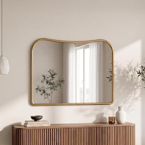 May include: A wall-mounted mirror featuring a thin, gold-toned metal frame with a distinctively curved, wavy top edge. The rectangular mirror reflects a bright interior space with white curtains and a small potted plant, creating a clear reflection.