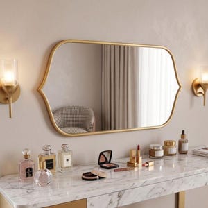 May include: A horizontal wall mirror with a gold frame featuring rounded corners and pointed sides, flanked by two gold wall sconces. The mirror reflects a room with a houndstooth chair and curtains, positioned above a white marble vanity table.