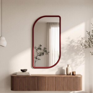 May include: A wall-mounted mirror with a deep red, rounded frame featuring a unique asymmetrical shape. The mirror is positioned above a long, horizontal, wall-mounted wooden console table with a vertical fluted texture along its front surface.
