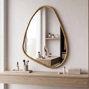 May include: A wall-mounted mirror with an irregular, organic, pebble-shaped frame finished in a smooth metallic gold. The reflective surface shows a clear view of a wooden vanity table, makeup brushes, and various small personal care items.