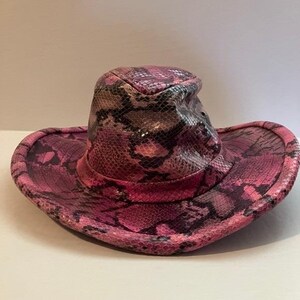 May include: A cowboy hat featuring a vibrant pink and black snakeskin pattern. The hat has a structured crown with a slight indentation at the top and a wide, curved brim. The material displays a textured, reptilian scale design throughout.