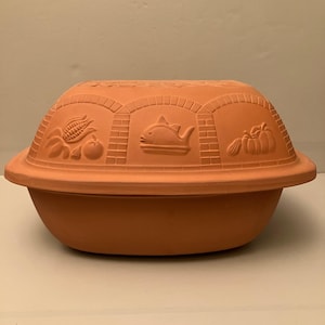 May include: A terracotta clay baker with a lid featuring embossed designs of corn, a tomato, a fish, and a pumpkin. The lid has a brick-patterned arch motif surrounding each food item, and the base is a smooth, deep, oval-shaped bowl.