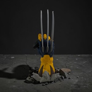 May include: A yellow and dark blue sculpted hand figurine featuring three long, sharp silver-colored claws extending upward. The base of the hand is encased in a yellow gauntlet, emerging from a cracked, gray, rocky-textured ground surface.