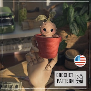 May include: A small, beige, round crocheted creature with two green leaves on top, black eyes, and pink cheeks, sitting inside a textured, terracotta-colored crocheted pot. The item is presented as a digital crochet pattern in PDF format.