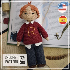 Pode incluir: Uma boneca feita à mão em crochê com cabelo ruivo, sardas e olhos pretos. A boneca veste um suéter bordô de manga comprida com uma letra R amarela bordada, uma camisa branca com gola por baixo, calças cinza-escuras e sapatos marrons.