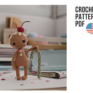 May include: A brown crocheted gingerbread doll with a white icing-style head topping, a red cherry on top, and two small buttons on its chest. The doll holds a green and white striped candy cane. Text reads CROCHET PATTERN PDF with a US flag icon.