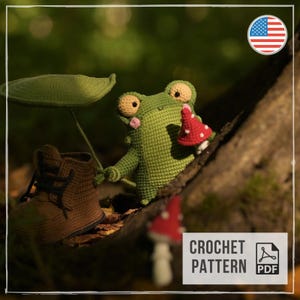 May include: A green crocheted frog toy holding a small red and white mushroom, standing next to a brown crocheted boot and a green leaf. The frog has large eyes and pink cheeks. The text CROCHET PATTERN PDF is visible in the corner.