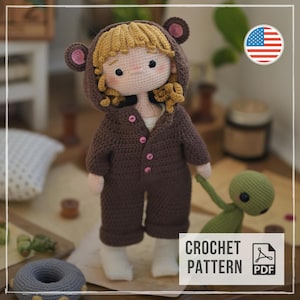 May include: A handmade crochet doll with curly blonde hair wearing a dark brown hooded bear onesie with pink buttons. The doll has a light skin tone, rosy cheeks, and black eyes. A small green crochet creature is held in one hand of the doll.