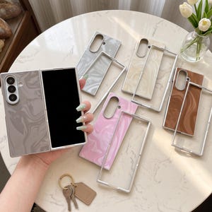 May include: A collection of foldable phone cases featuring a marbled pattern in gray, beige, pink, and brown. Each case consists of two parts with a silver-toned frame and a smooth, glossy finish. One case is shown held up, displaying the camera cutout.