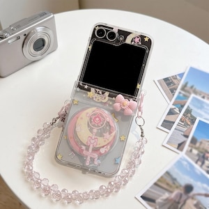 May include: A clear flip phone case featuring a Sailor Moon-themed design with a crescent moon, a star-topped wand, and a small pink bow. The case includes a detachable beaded wrist strap made of translucent, faceted beads and a camera cutout.