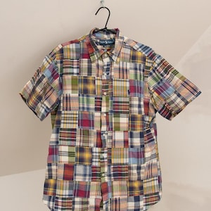 May include: A short-sleeve button-down shirt featuring a multi-coloured patchwork plaid pattern in shades of red, blue, yellow, green, and white. The shirt has a standard collar, a front button placket, and a small dark logo on the left chest.