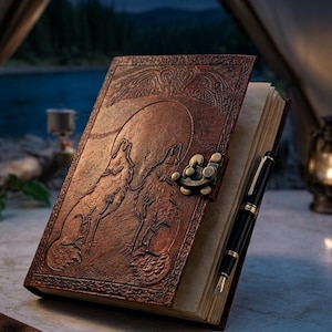 May include: A brown leather-bound journal featuring an embossed design of two wolves howling at a large moon on the front cover. The journal includes a brass-colored metal latch closure and off-white pages, with a black and gold fountain pen.
