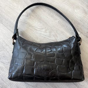 Könnte beinhalten: Eine schwarze Schultertasche mit glänzender Krokodilprägung. Die Tasche verfügt über einen einzelnen geschwungenen Trageriemen, der mit messingfarbenen Metallringen befestigt ist, sowie ein ovales Emblem mit Baum-Logo auf der Vorderseite.