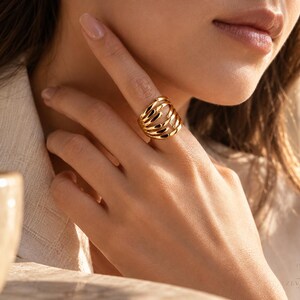 Puede incluir: Un anillo de tono dorado con un diseño de múltiples bandas que consta de cinco hilos de metal paralelos, curvos y pulidos que se envuelven alrededor del dedo. El anillo tiene una superficie lisa y reflectante y una estructura ancha y abierta.
