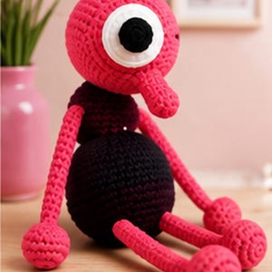 May include: A handmade crochet doll featuring a round hot pink head with a single large white and black eye, a small pink nose, a dark purple torso, and long hot pink limbs ending in round feet. The doll is constructed from yarn with visible stitches.