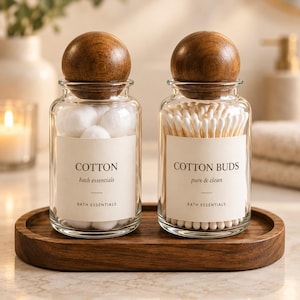 May include: Two clear glass jars with spherical wooden lids, resting on an oval wooden tray. The left jar contains white cotton balls and a label reading COTTON bath essentials BATH ESSENTIALS. The right jar holds cotton buds and a label reading COTTON BUDS pure & clean BATH ESSENTIALS.