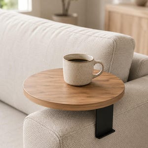 Könnte beinhalten: Ein rundes Sofa-Armlehnen-Tablett aus Holz mit einer schwarzen Metallklemme und einer hellbraunen Holzmaserung. Eine gesprenkelte beige Keramiktasse, gefüllt mit einer dunklen Flüssigkeit, steht mittig auf der flachen, runden Holzoberfläche.