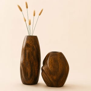 Op de afbeelding: Twee donkerbruine houten vazen met een gladde, gepolijste afwerking en zichtbare houtnerfpatronen. De hogere, cilindrische vaas bevat vijf dunne, lichtbruine gedroogde stengels met pluizige, ovale toppen, terwijl de kortere vaas rond is.