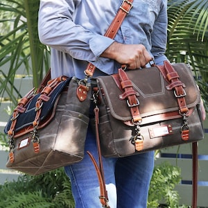 May include: Two brown canvas messenger bags with leather accents and straps. The bags have multiple pockets, buckles, and metal hardware. One bag has a dark blue flap. The bags are carried by a person wearing a light blue shirt and blue jeans.