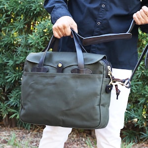 May include: A dark green canvas briefcase with a leather handle and strap. The briefcase has a flap closure and a zippered pocket on the front.